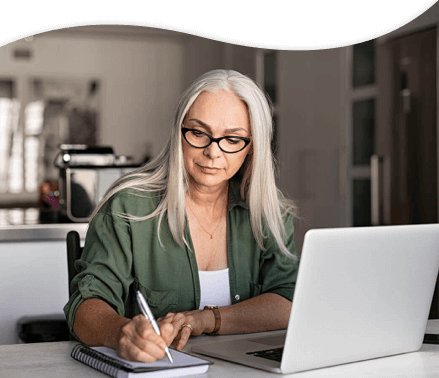 Woman sitting down at desk writing on notebook with laptop opened.